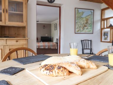 A dining table with croissants, bread, glasses of orange juice, wooden chairs, a wooden cupboard, and a framed map on the wall at Haulfryn in Bethesda