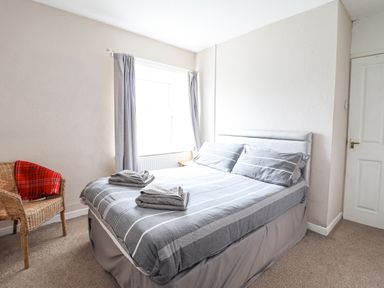 A bedroom with a double bed with grey striped bedding and two folded towels a window with grey curtains and a wicker chair with a red blanket at Haulfryn in Bethesda