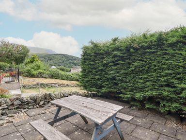 An outdoor patio area with a wooden picnic table and bench surrounded by stone walls and greenery at Haulfryn in Bethesda