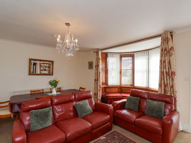A living room with red leather sofas a dining table with flowers and a bay window with vertical blinds at Mayfield Manor in Whitby