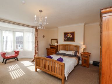 A bedroom with a wooden bed, wooden dresser, bedside table with lamp, red upholstered bench, and large bay window at Mayfield Manor in Whitby