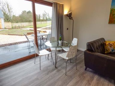 A dining area with a round glass table and four chairs next to sliding glass doors overlooking a garden and patio with outdoor furniture at Bramshurst in Upwey near Weymouth