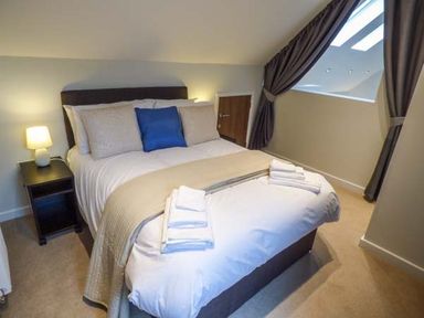 A bedroom with a bed with beige bedding and blue pillow a lamp on a nightstand and folded towels on the bed at Bramshurst in Upwey near Weymouth