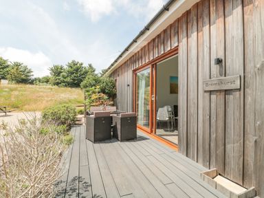 An outdoor wooden deck with table and chairs next to a building with sliding glass doors and a wooden sign reading Bramshurst at Bramshurst Upwey near Weymouth