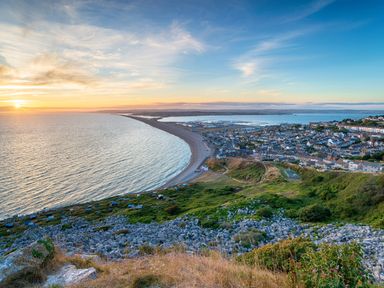 A coastal town with a curved beach at sunset with green hills and rocky terrain at Bramshurst Upwey near Weymouth
