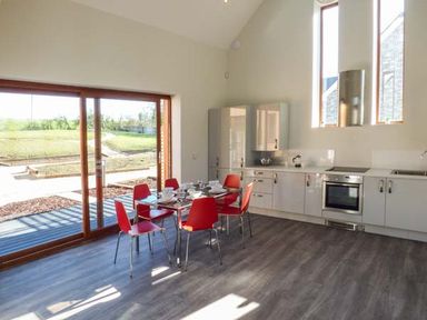 A kitchen with white cabinets a glass dining table with red chairs and large sliding glass doors at Casterbridge in Upwey near Weymouth