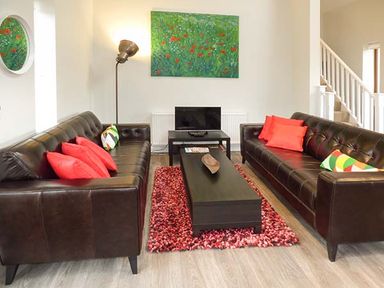 A living room with two dark brown leather sofas with red and multicolored pillows a black coffee table on a red rug a floor lamp and a green floral painting at Casterbridge Upwey near Weymouth