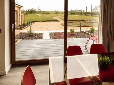 A dining area with red chairs and glass table near a large window overlooking a garden and patio at Casterbridge Upwey near Weymouth