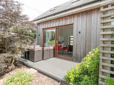 A wooden deck with outdoor seating and a table outside a house with sliding glass doors showing an interior dining area at Casterbridge in Upwey near Weymouth