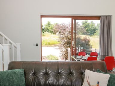 A living room with a dark brown leather sofa and cushions near a large glass sliding door with red chairs at Casterbridge Upwey near Weymouth