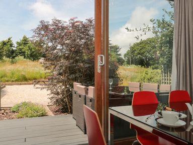A dining area with red chairs and a glass table next to a sliding door opening to a garden with outdoor seating at Casterbridge Upwey near Weymouth