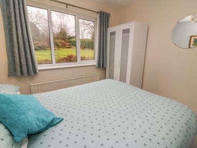 A bedroom with a bed with patterned bedding a blue pillow a white wardrobe a round mirror and a window with curtains showing a garden at Trederwen Rhayader