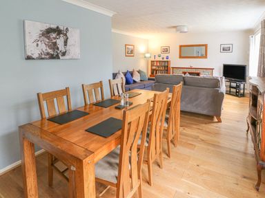 A dining area with a wooden table and six chairs next to a living room with sofas a TV and a bookshelf at Trederwen in Rhayader