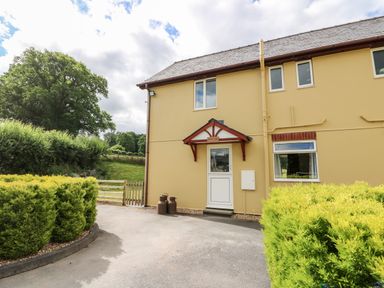 The exterior of a yellow two-story house with a white door and red canopy surrounded by green bushes at Red Kite Cottage in Howey near Llandrindod Wells