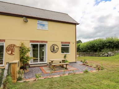 An outdoor patio area with a wooden picnic table and benches outside a house with yellow walls and two wall decorations at Red Kite Cottage in Howey near Llandrindod Wells