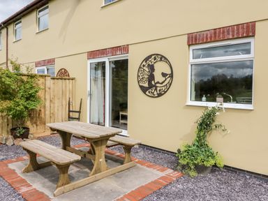 An outdoor patio with a wooden picnic table and benches next to a yellow building with a sliding glass door and a window with a potted plant below and a metal wall decoration at Red Kite Cottage in Howey near Llandrindod Wells