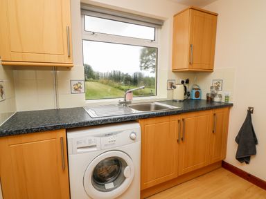A kitchen with wooden cabinets a washing machine a sink and a window showing a garden at Red Kite Cottage in Howey near Llandrindod Wells