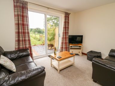 A living room with two brown leather sofas a wooden coffee table a television on a stand and glass sliding doors with red plaid curtains at Red Kite Cottage in Howey near Llandrindod Wells