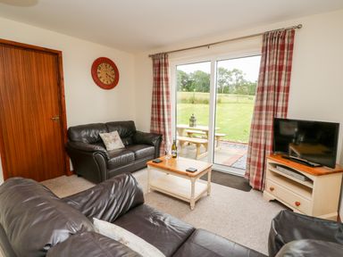 A living room with dark leather sofas a coffee table and a television near sliding glass doors with plaid curtains at Red Kite Cottage in Howey near Llandrindod Wells