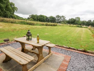 A wooden picnic table with a lantern wine bottle and two glasses on a patio overlooking a fenced grassy field with sheep at Red Kite Cottage in Howey near Llandrindod Wells