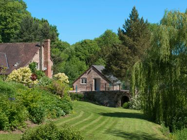 A garden with lawn and shrubs next to stone buildings and trees at Mill Cottage Hanley William near Tenbury Wells