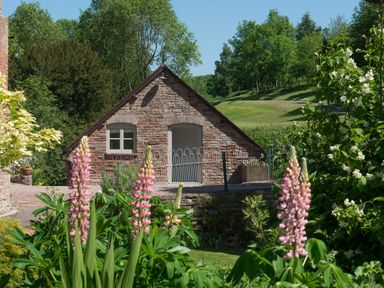 A stone building with a fenced entrance surrounded by greenery and pink flowers at Mill Cottage in Hanley William near Tenbury Wells