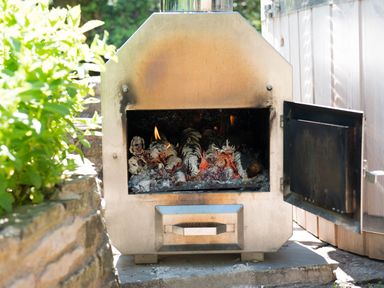 Outdoor metal fire pit with burning logs next to a stone wall and greenery at Mill Cottage in Hanley William near Tenbury Wells