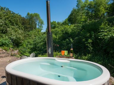 An outdoor hot tub with two colored glasses and a bottle on a wooden tray in a garden area at Mill Cottage in Hanley William near Tenbury Wells