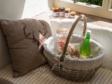 A wicker basket with granola, baked goods, and a green bottle next to a cushion with a stag design by a window at Mill Cottage in Hanley William near Tenbury Wells