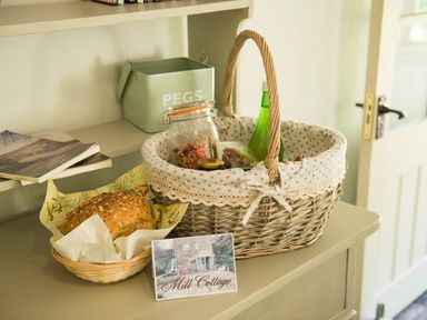A wicker basket with a jar of granola and a green bottle next to a basket with bread on a dresser at Mill Cottage Hanley William near Tenbury Wells