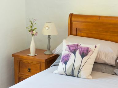 A bed with a wooden headboard a pillow with purple flowers and a wooden bedside table with a lamp and vase at Mill Cottage in Hanley William near Tenbury Wells
