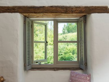 A window with grey shutters open to a view of green trees and bushes at Mill Cottage in Hanley William near Tenbury Wells