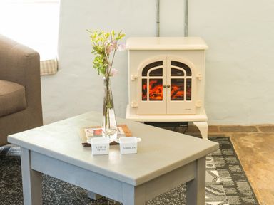 A room with a gray coffee table holding a glass vase with flowers and small containers near a beige stove and a brown sofa at Mill Cottage in Hanley William near Tenbury Wells