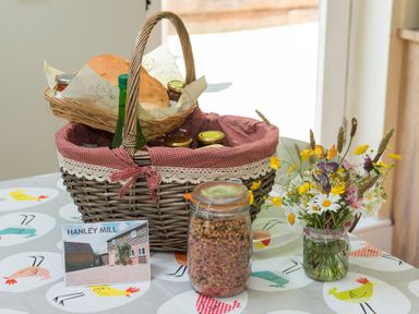 A basket with bread jars and a bottle on a table with a jar of granola flowers and a postcard at Stable Cottage in Hanley William near Tenbury Wells