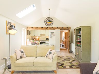 A living room with a beige sofa and pillows facing a kitchen with white cabinets and a wooden beam at Stable Cottage Hanley William near Tenbury Wells
