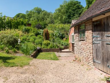 A garden with shrubs and plants next to a stone building with wooden doors at Stable Cottage in Hanley William near Tenbury Wells