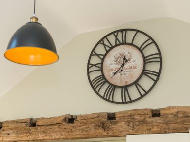 A wall clock with roman numerals above a wooden beam and a black hanging lamp at Stable Cottage Hanley William near Tenbury Wells