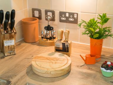 A kitchen countertop with a knife block a corkscrew set a cheese board cheese knives a plant in an orange pot an orange watering can and a cupcake container at Stable Cottage in Hanley William near Tenbury Wells