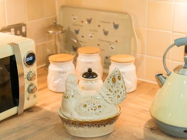 A kitchen countertop with a ceramic hen container a yellow kettle a microwave and white canisters with wooden lids at Stable Cottage Hanley William near Tenbury Wells