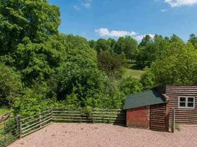 An outdoor scene with a wooden fence gravel ground and a small wooden and brick shed surrounded by trees at Stable Cottage in Hanley William near Tenbury Wells