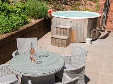 An outdoor patio with a round glass table and four wicker chairs next to a wooden hot tub at Stable Cottage in Hanley William near Tenbury Wells