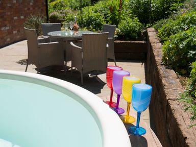 An outdoor patio with a round table and four wicker chairs a bottle and glasses on the table and colorful plastic wine glasses on the edge of a round hot tub at Stable Cottage Hanley William near Tenbury Wells