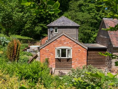 A brick and wood house surrounded by green trees and plants at Stable Cottage Hanley William near Tenbury Wells