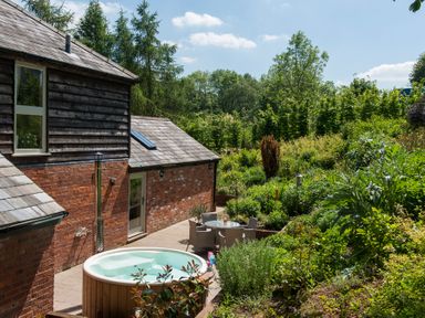 An outdoor patio area with a hot tub and a round glass table with chairs next to a brick and wood house surrounded by greenery at Stable Cottage in Hanley William near Tenbury Wells