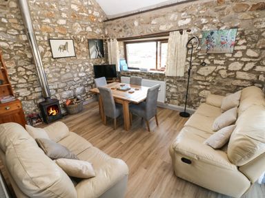 A living room with beige sofas a wooden dining table with grey chairs a wood burning stove and stone walls at Corner Cottage in Broad Haven