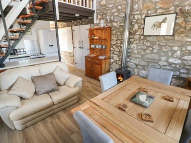 An open plan living area with a wooden dining table four chairs a cream sofa with cushions a wooden cabinet a wood burning stove and kitchen appliances at Corner Cottage in Broad Haven