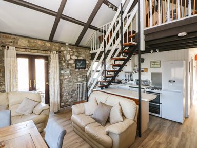 A living area with beige sofas wooden table and chairs kitchen area with white appliances and staircase leading to loft at Corner Cottage in Broad Haven