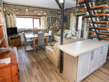 A living room with stone walls a dining table with chairs a TV a cream sofa and a staircase at Corner Cottage in Broad Haven