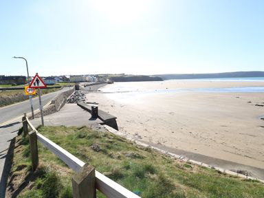 A coastal road with a pedestrian warning sign next to a sandy beach and houses in the distance at Corner Cottage in Broad Haven