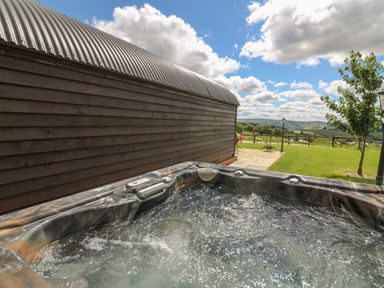 An outdoor hot tub next to a wooden building with a view of a grassy field and hills at Valley View in Rhayader